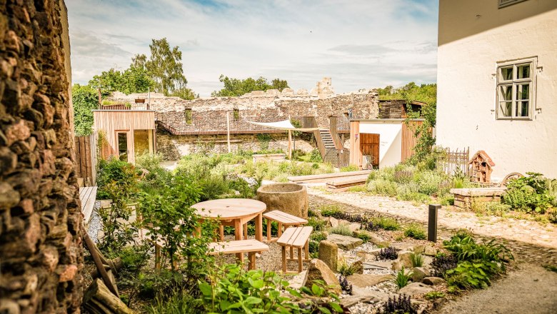 Ein Garten mit Holztischen und B&auml;nken, umgeben von alten Steinmauern und Pflanzen, unter einem blauen Himmel.