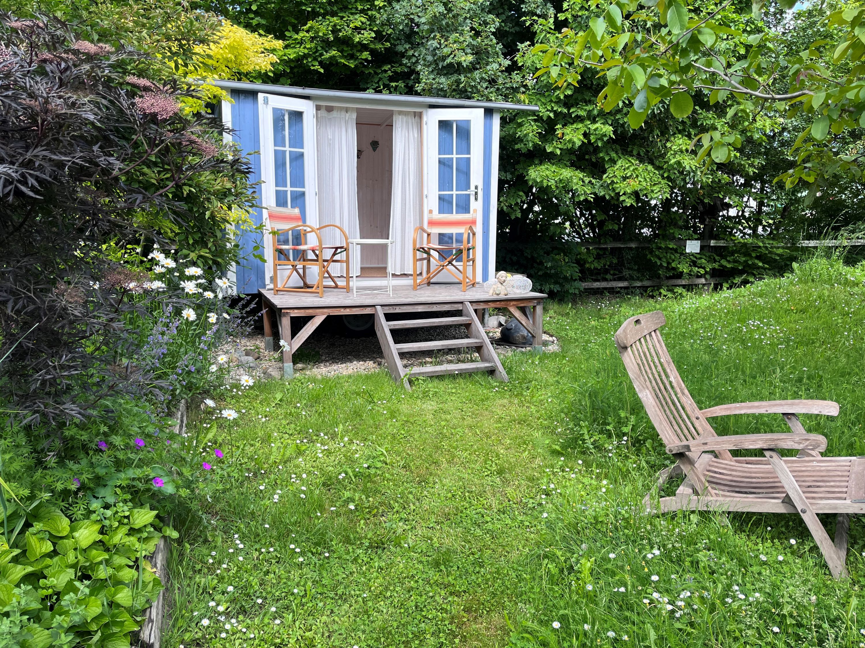 Ein blauer Gartenpavillon mit zwei Stühlen auf einer Holzveranda, umgeben von einer grünen Wiese und bunten Blumen.