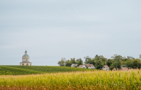Landschaft mit einem runden Geb&auml;ude und mehreren kleinen H&auml;usern am Waldrand.