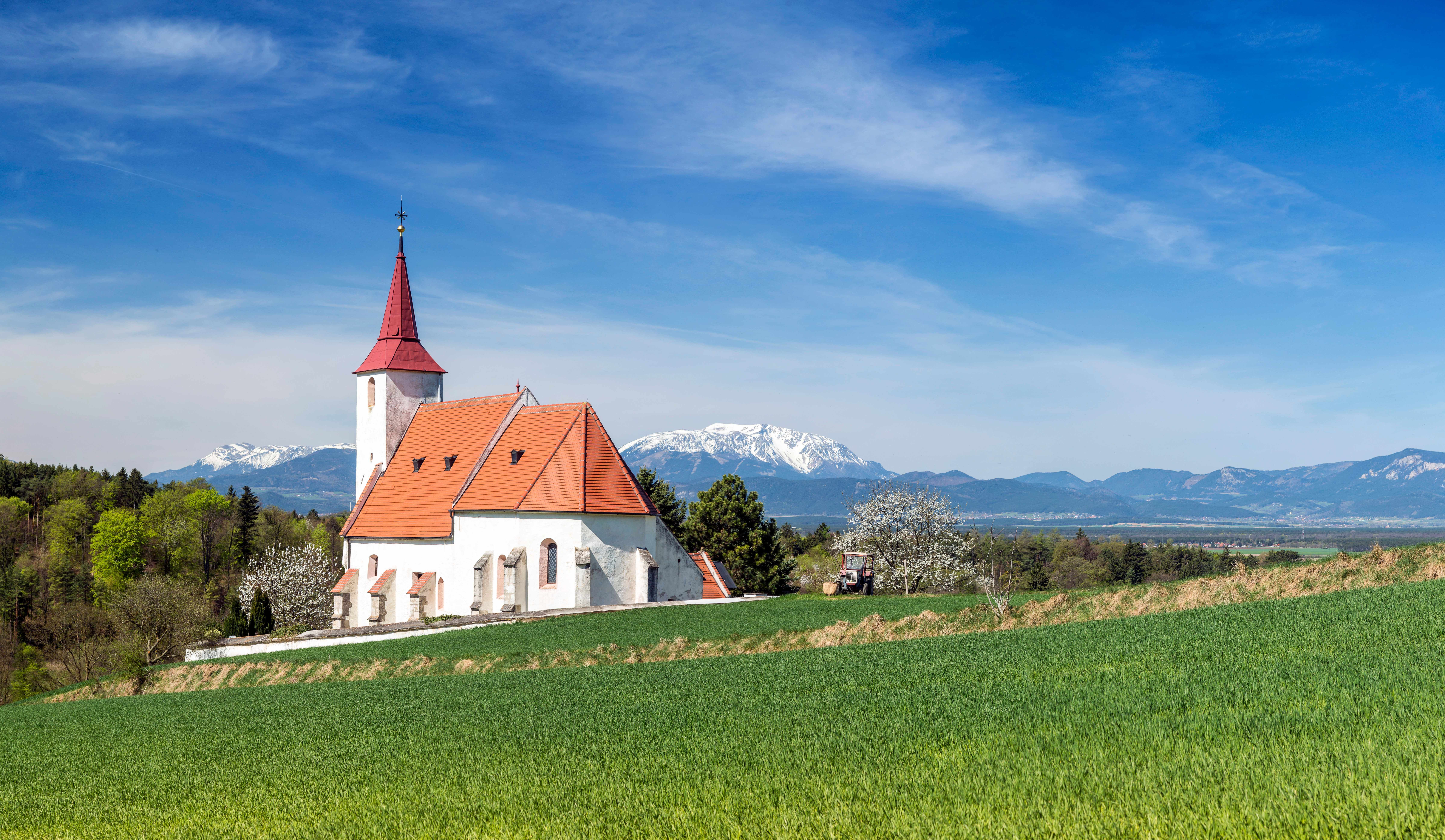 Weiße Pfarrkirche Ofenbach mit rotem Dach und Kirchturm vor einer Bergkulisse mit grünem Feld im Vordergrund.