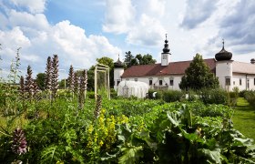Ein Klostergeb&auml;ude mit Zwiebelt&uuml;rmen hinter einem &uuml;ppigen Garten unter blauem Himmel.