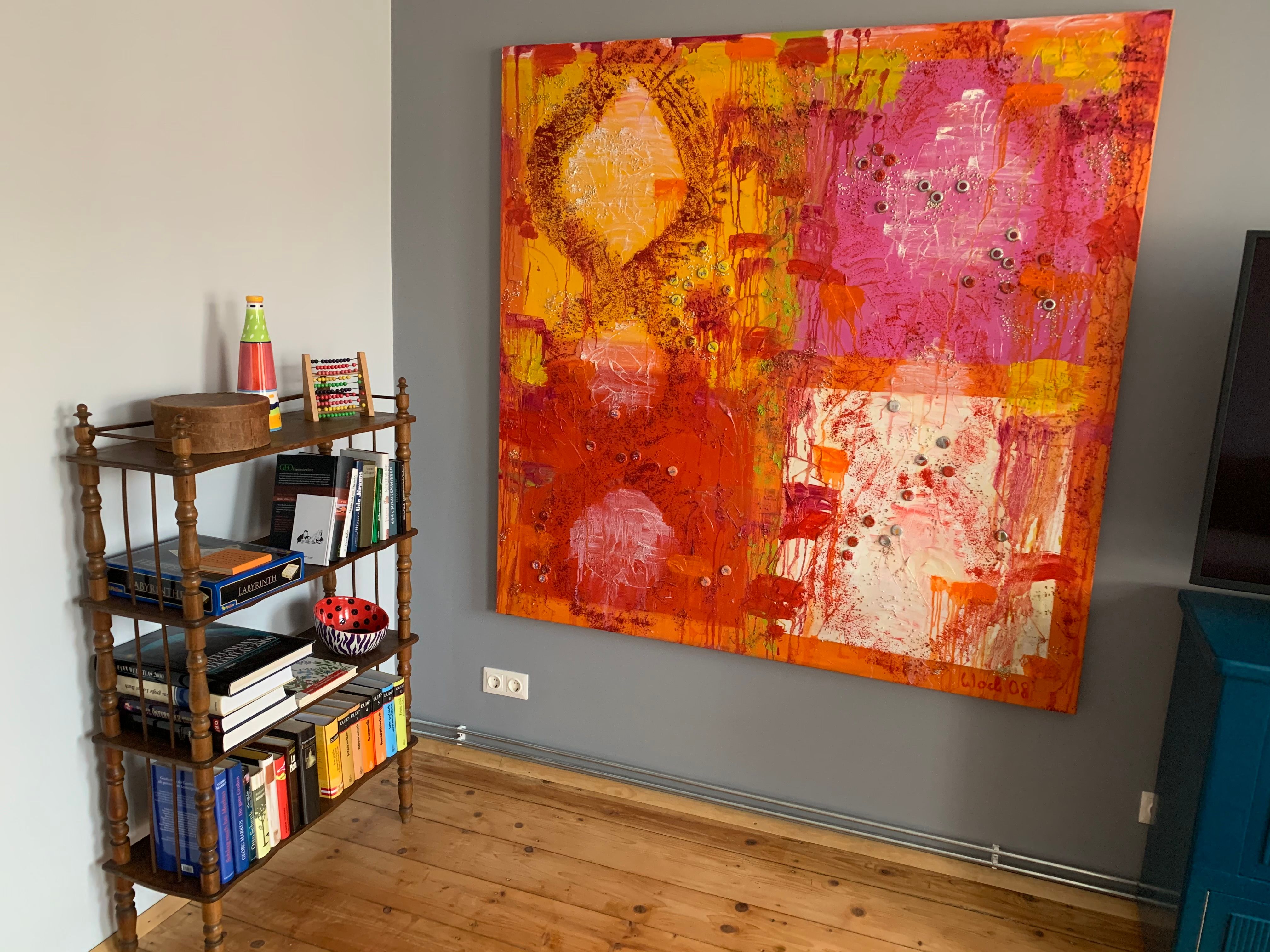 Ein kleines Bücherregal mit Büchern und Dekorationen neben einem großen, bunten Gemälde an der Wand.