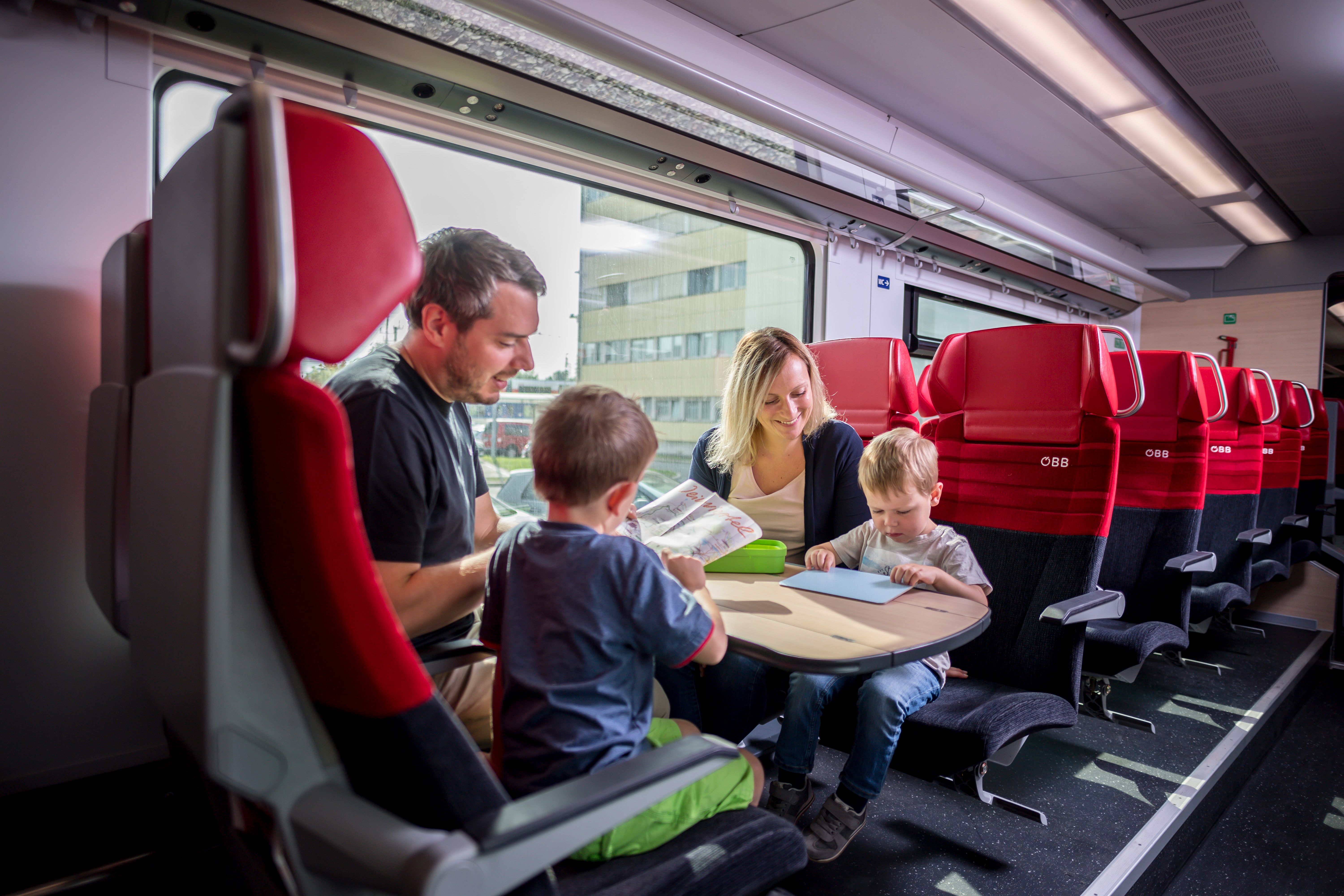 Eine Familie sitzt in einem Zugabteil mit roten Sitzen und spielt zusammen an einem Tisch.