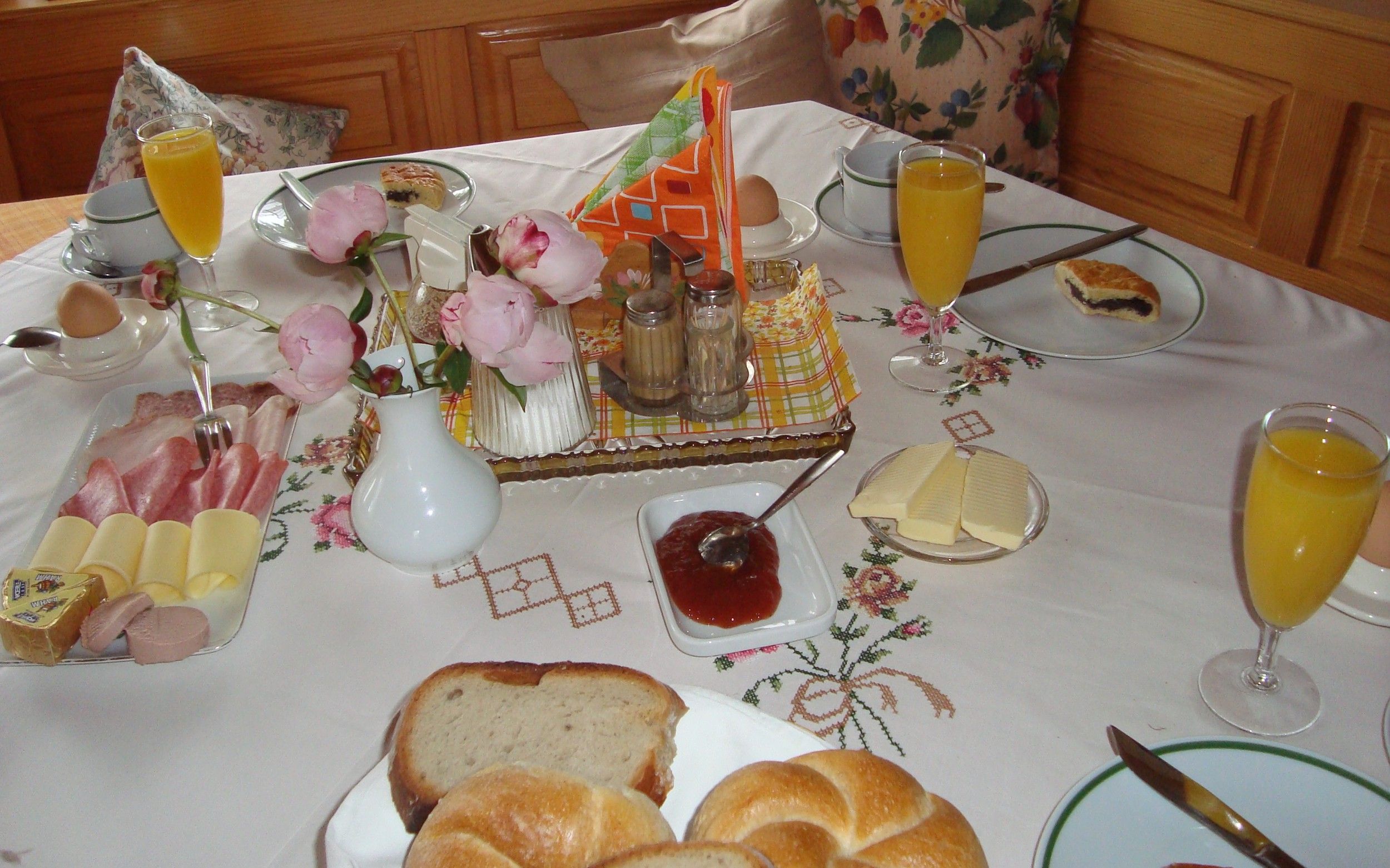 Ein gedeckter Frühstückstisch mit Brötchen, Aufschnitt, Käse, Marmelade, Orangensaft und Blumen.