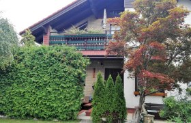 Au&szlig;enansicht eines Hauses mit Balkon, umgeben von B&auml;umen und Str&auml;uchern.