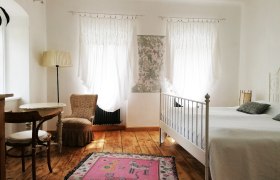 Helles Zimmer mit Holzboden, wei&szlig;em Bett, zwei Fenstern mit Vorh&auml;ngen, einem Tisch mit Stuhl und einem rosa Teppich.