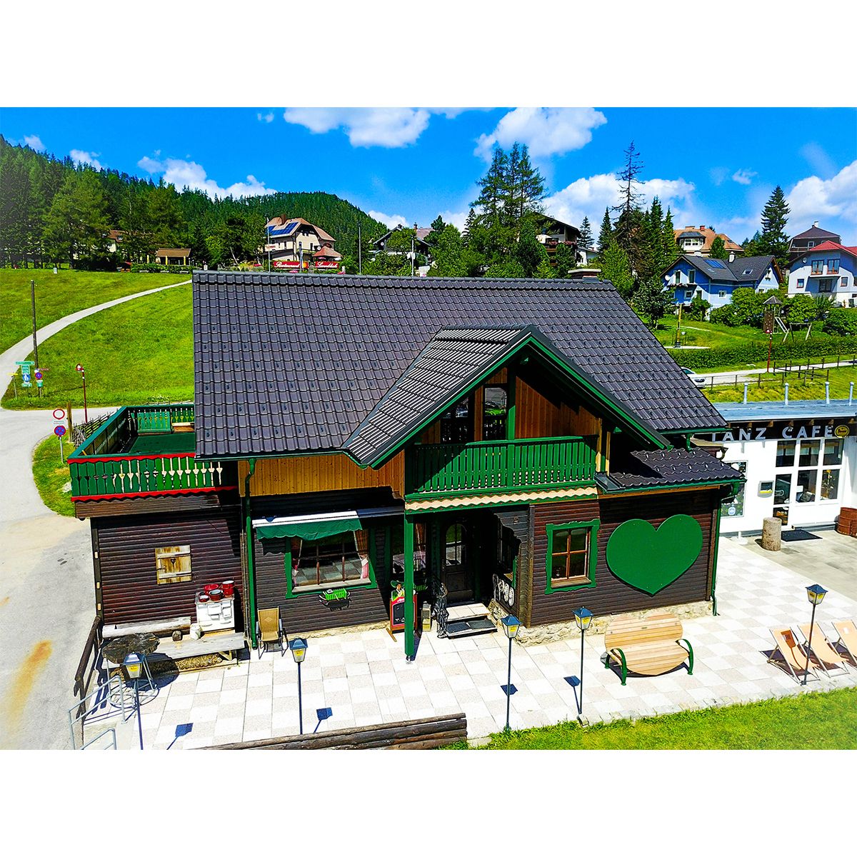 Ein traditionelles Holzhaus mit grünem Herzmotiv und Terrasse im Sommer, umgeben von grünen Hügeln und blauem Himmel.