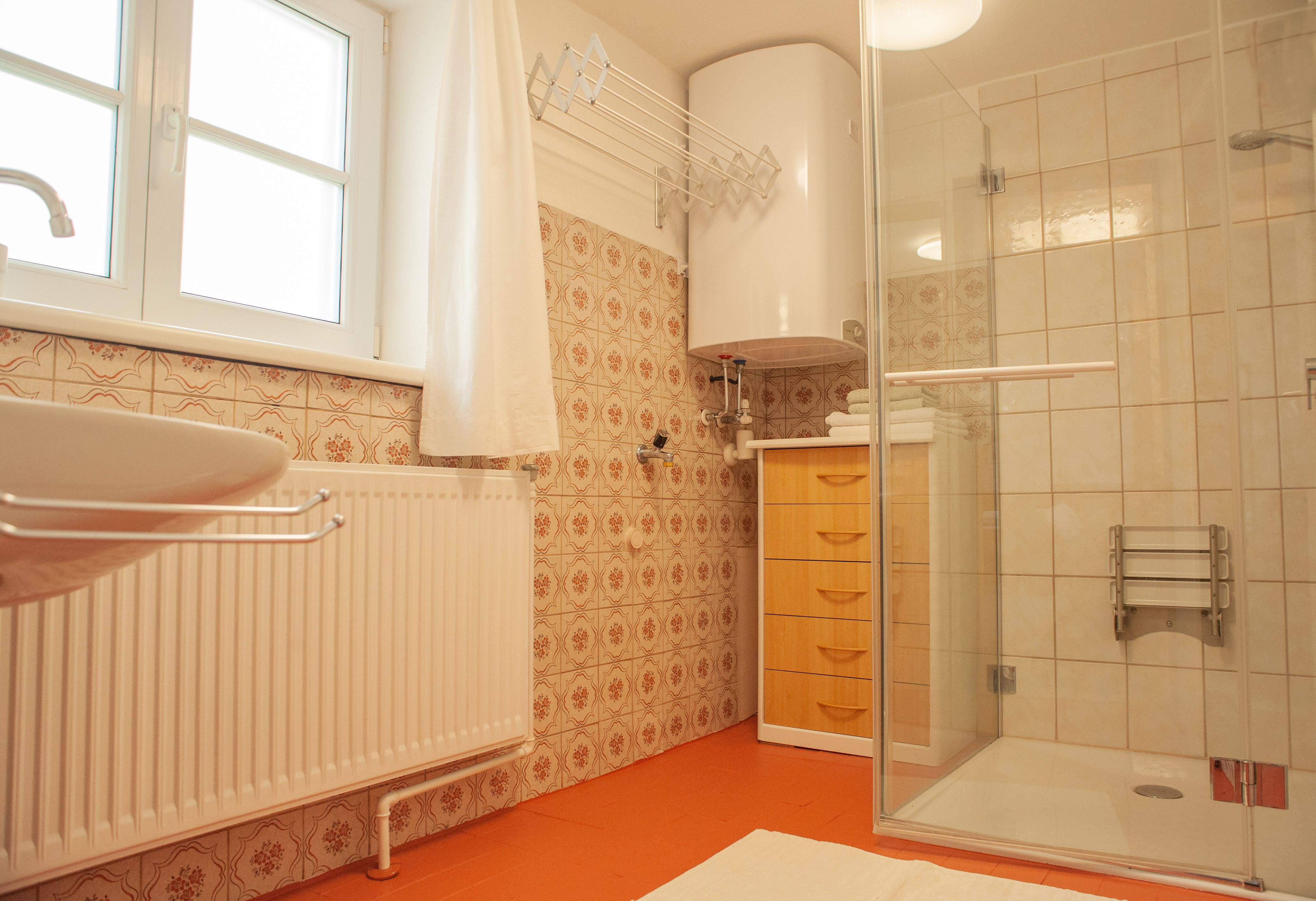 Ein Badezimmer mit Dusche, Waschbecken, Heizkörper und Holzschrank. Die Wände sind mit gemusterten Fliesen verkleidet.