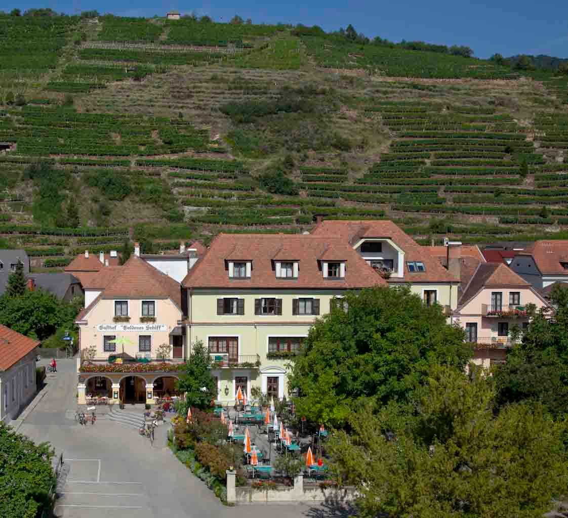 Außenansicht eines Gasthauses mit Terrasse und Weinbergen im Hintergrund.