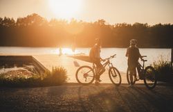 Zwei Radfahrerinnen mit Helmen stehen mit ihren Rädern bei tief stehender Sonne am Ufer des Viehofner Sees in St. Pölten.