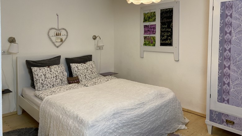 Gem&uuml;tliches Doppelzimmer mit wei&szlig;em Bett, dekorativem Herzregal und lila Schrank.