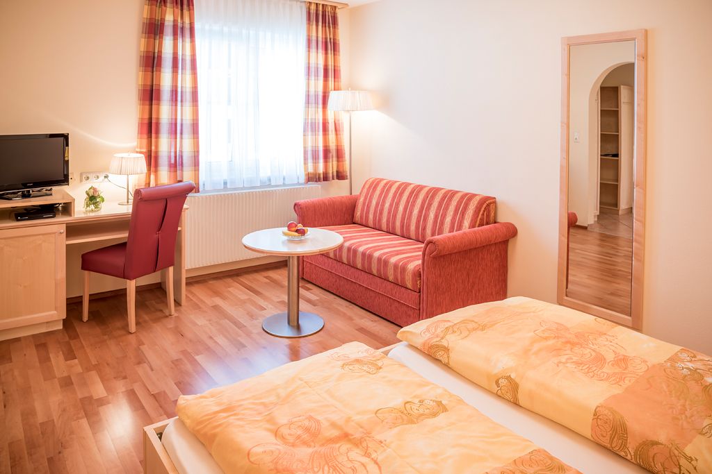 Gemütliches Gästezimmer mit Doppelbett, rotem Sofa, Schreibtisch, Fernseher und großem Spiegel.