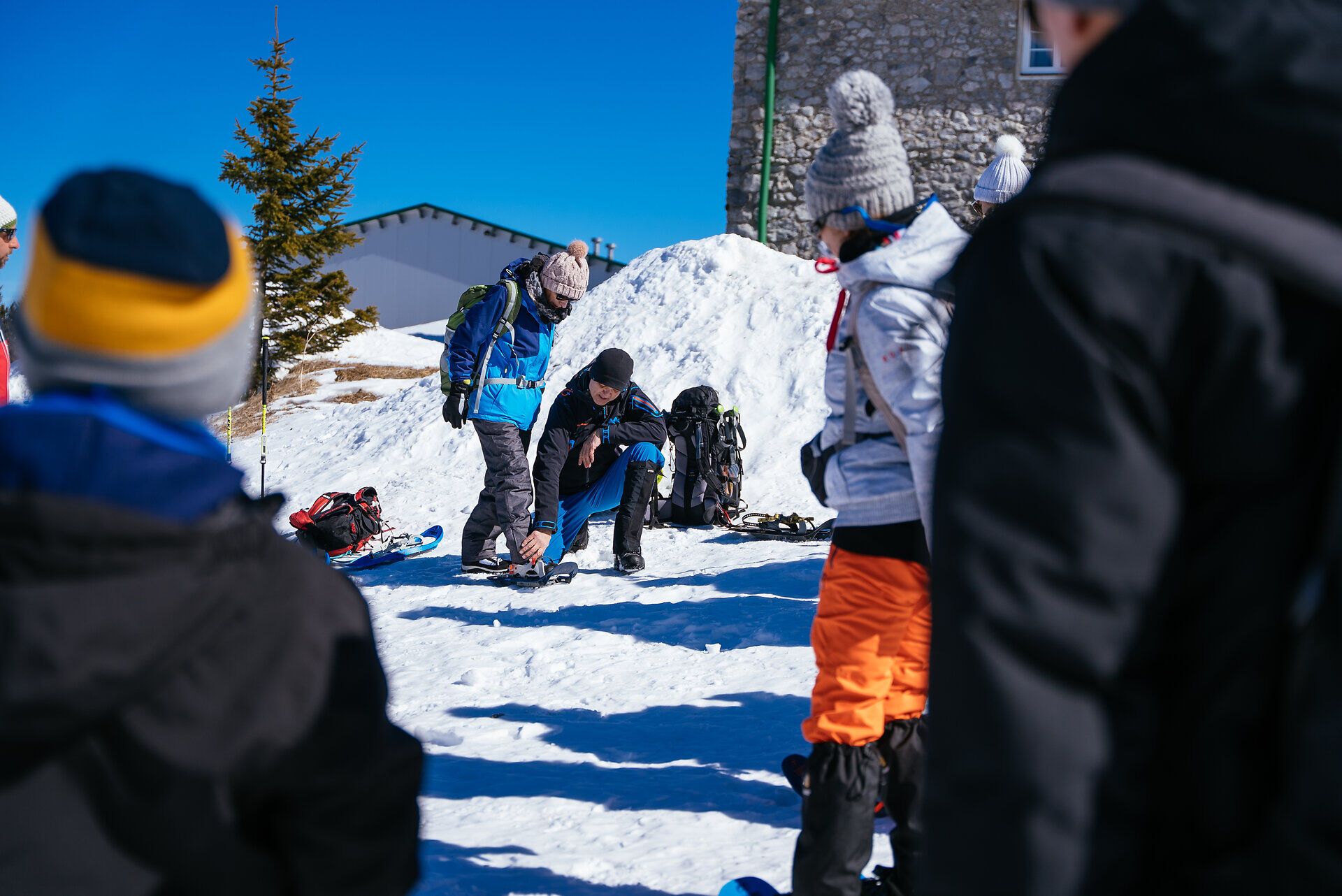 Gruppe von Menschen mit Schneeschuhen wandert auf verschneiter Fläche vor Nadelbäumen unter blauem Himmel