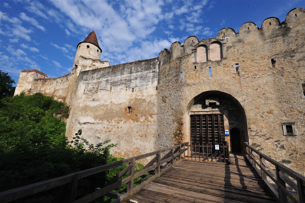 Eingangstor einer alten Burg mit Holztor und Zinnenmauer, blauer Himmel im Hintergrund.