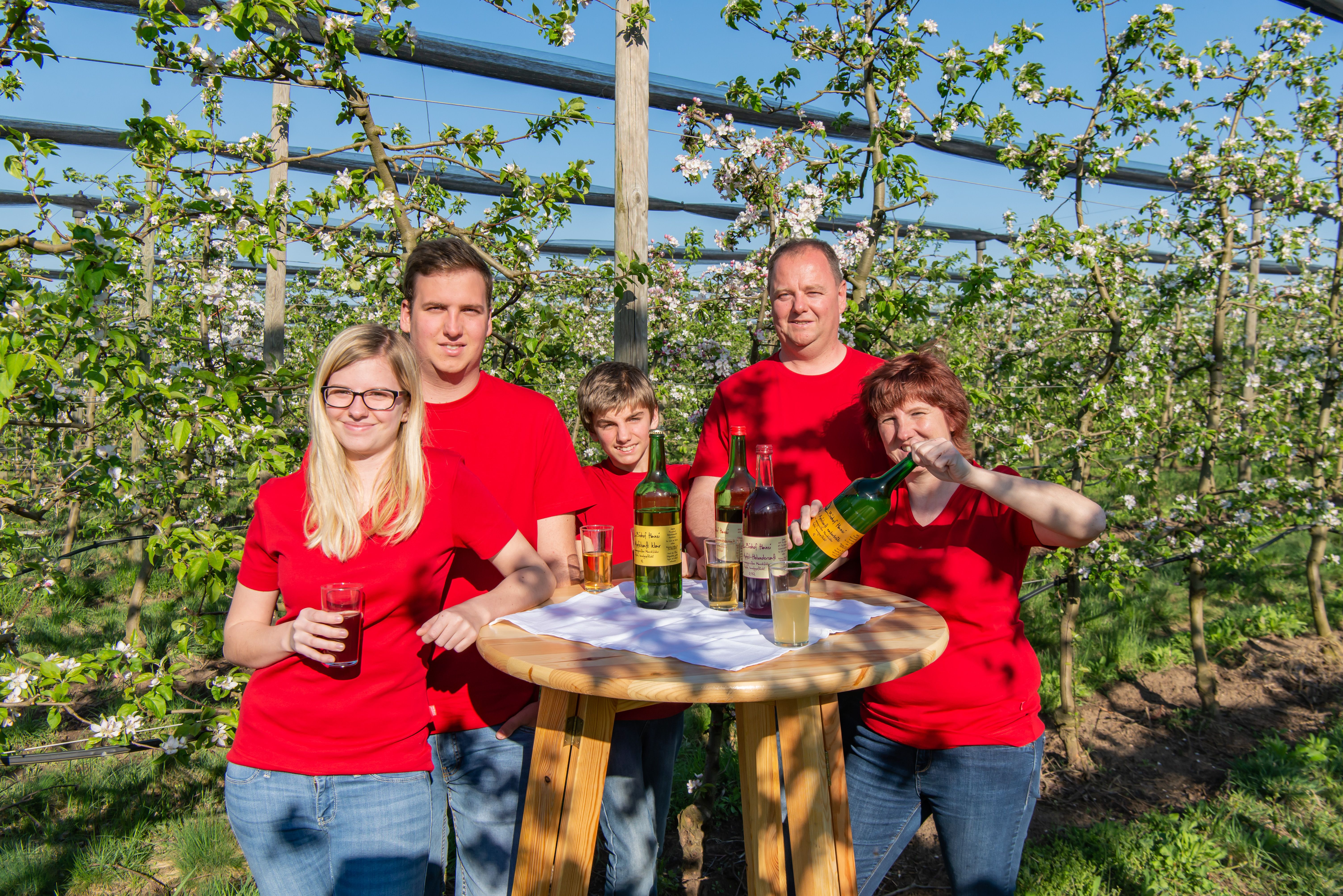 Eine Familie in roten T-Shirts steht an einem Holztisch mit Getränken in einem blühenden Obstgarten.