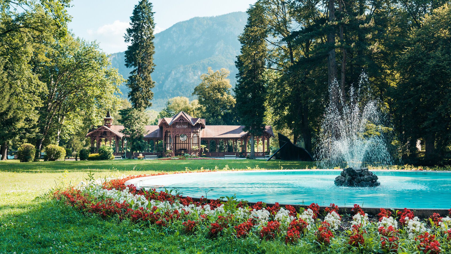 Parkanlage mit Springbrunnen, Blumenbeet und historischem Pavillon vor Bergkulisse an einem sonnigen Tag.