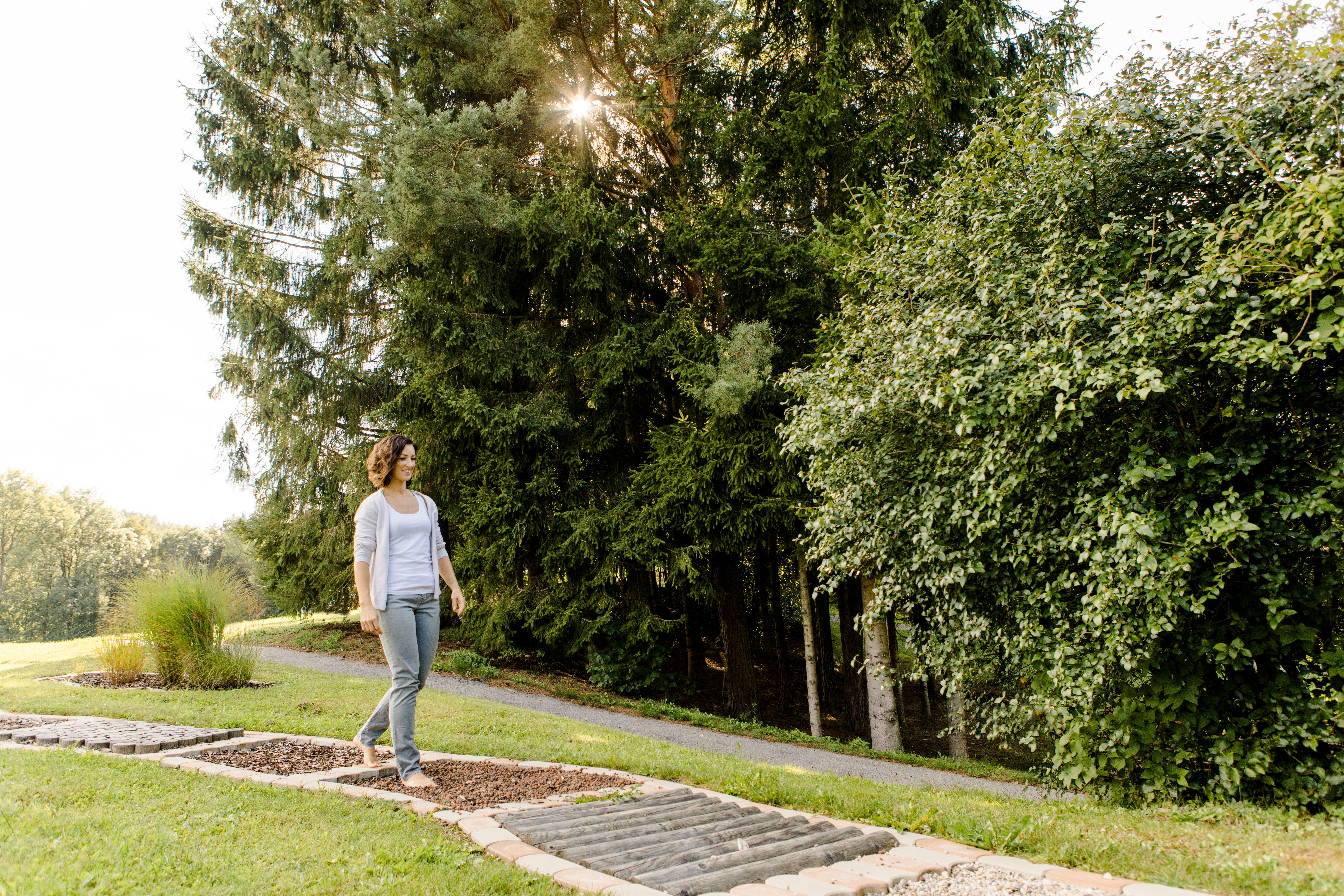 Eine Frau geht barfuß auf einem Pfad im Kurpark Bad Schönau, umgeben von Bäumen und Gras.
