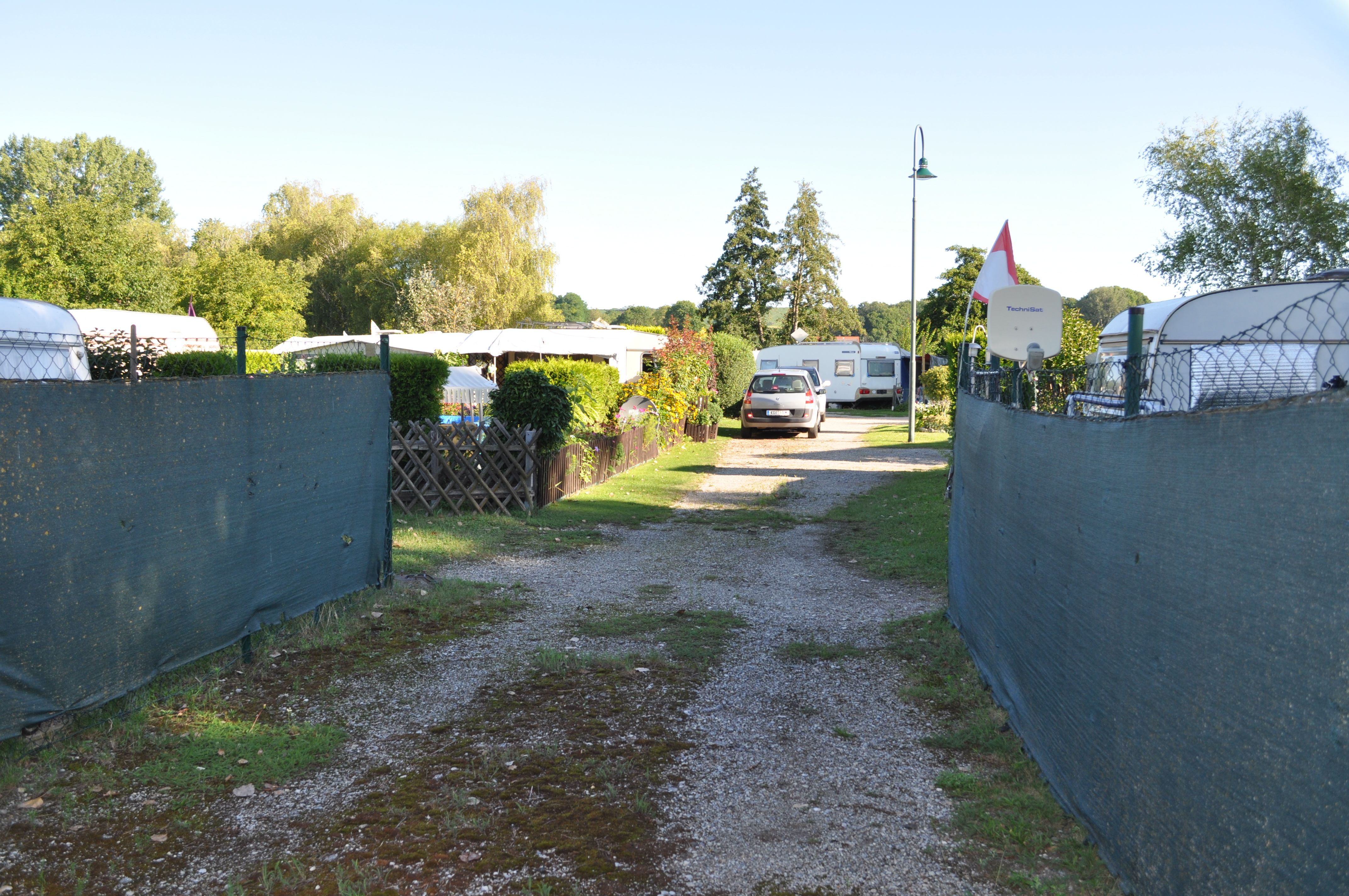 Ein Campingplatz mit Wohnwagen und einem Auto auf einem Schotterweg, umgeben von Bäumen und Zäunen.