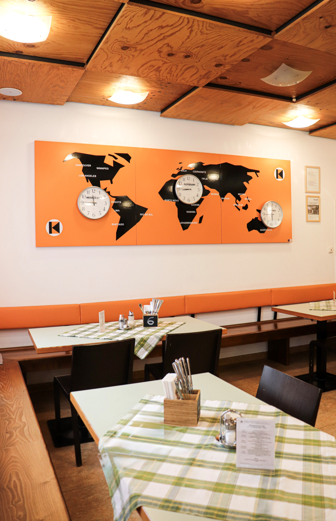 Innenraum des Kolpinghauses mit Holzbalkendecke, orangefarbener Weltkarten-Wand und Tischen mit karierten Tischdecken.