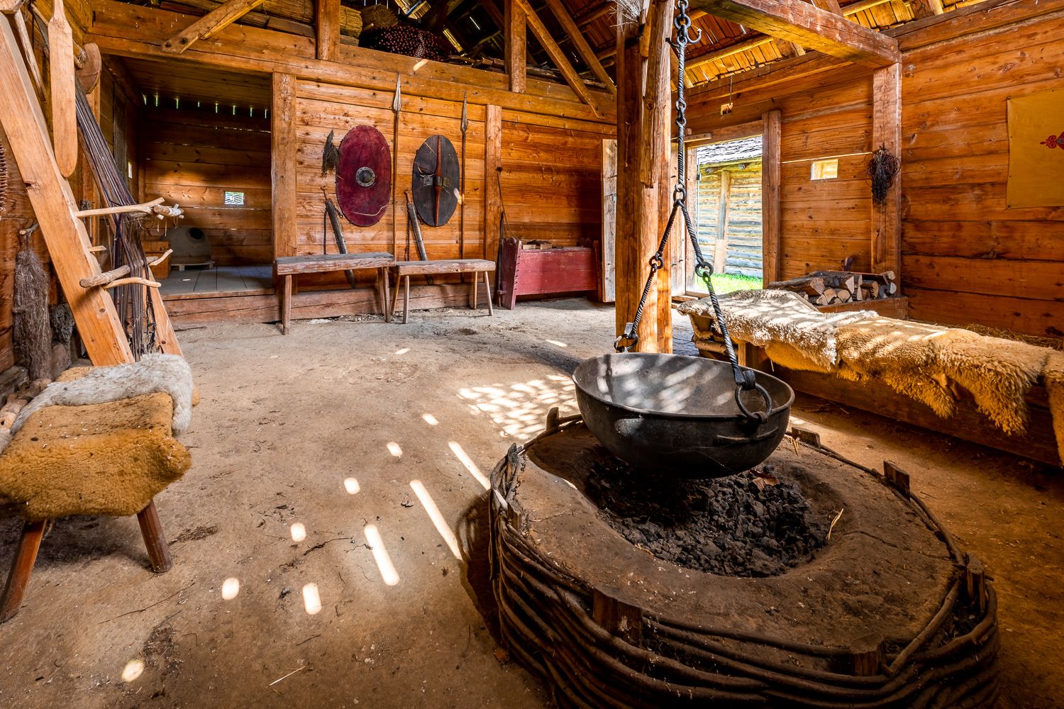 Innenansicht eines rekonstruierten keltischen Hauses mit Holzwänden, einem großen Kessel über einer Feuerstelle und Schilden an der Wand.