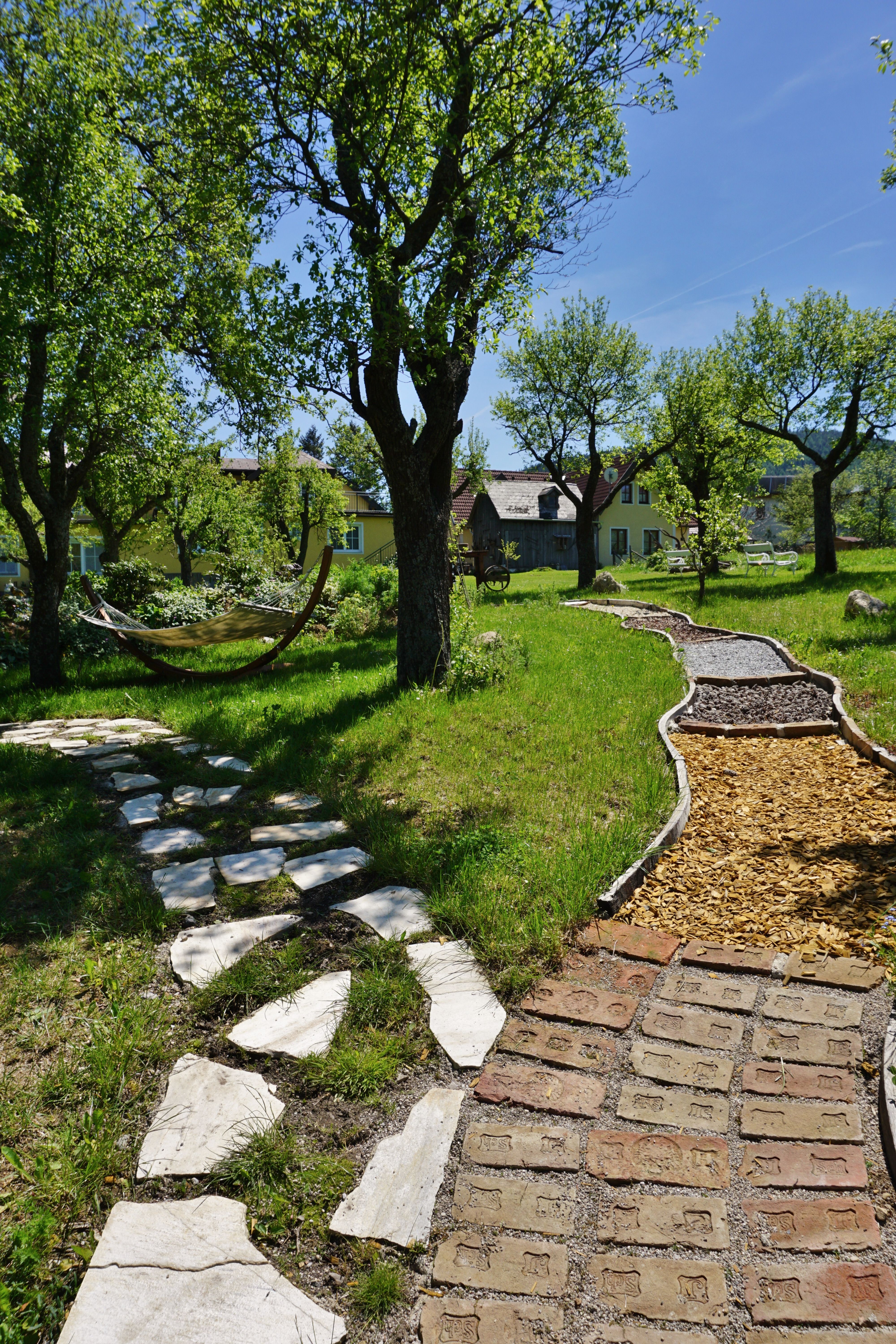 Ein Barfußweg mit verschiedenen Materialien in einem grünen Garten mit Bäumen und einer Hängematte.