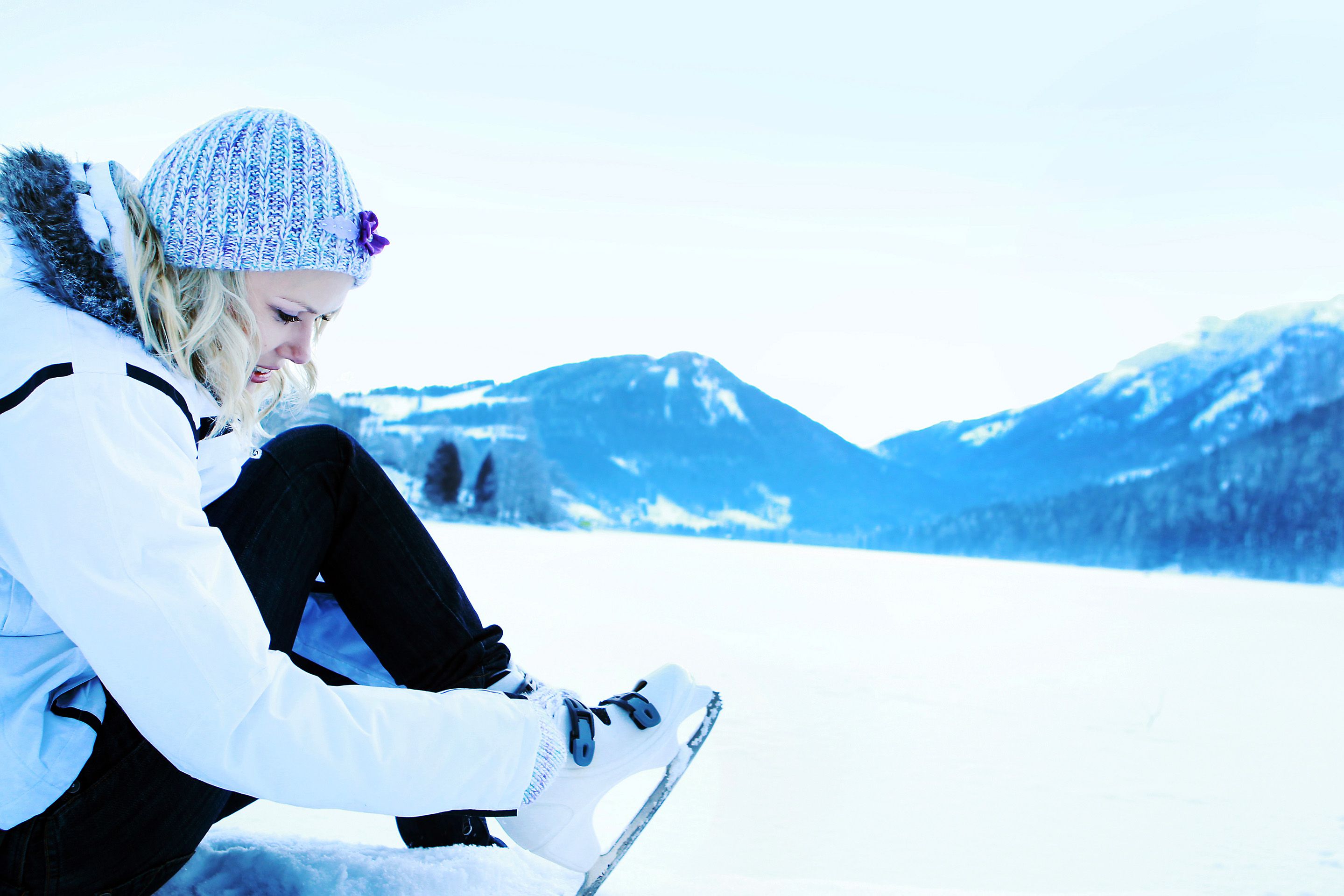 Person zieht Schlittschuhe an einem verschneiten See an, umgeben von Bergen.