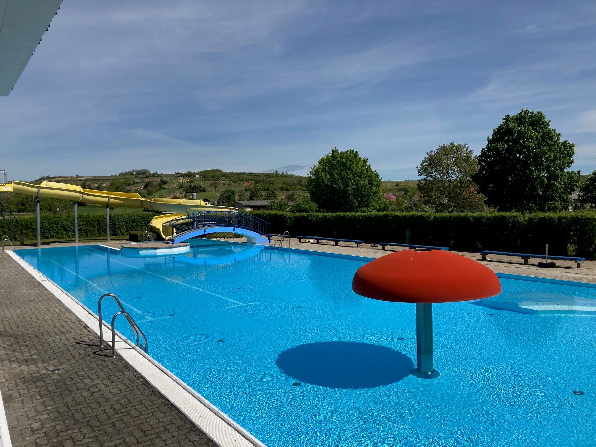 Ein Freibad mit einer gelben Wasserrutsche, einem blauen Becken und einem roten Wasserspiel in Form eines Pilzes.