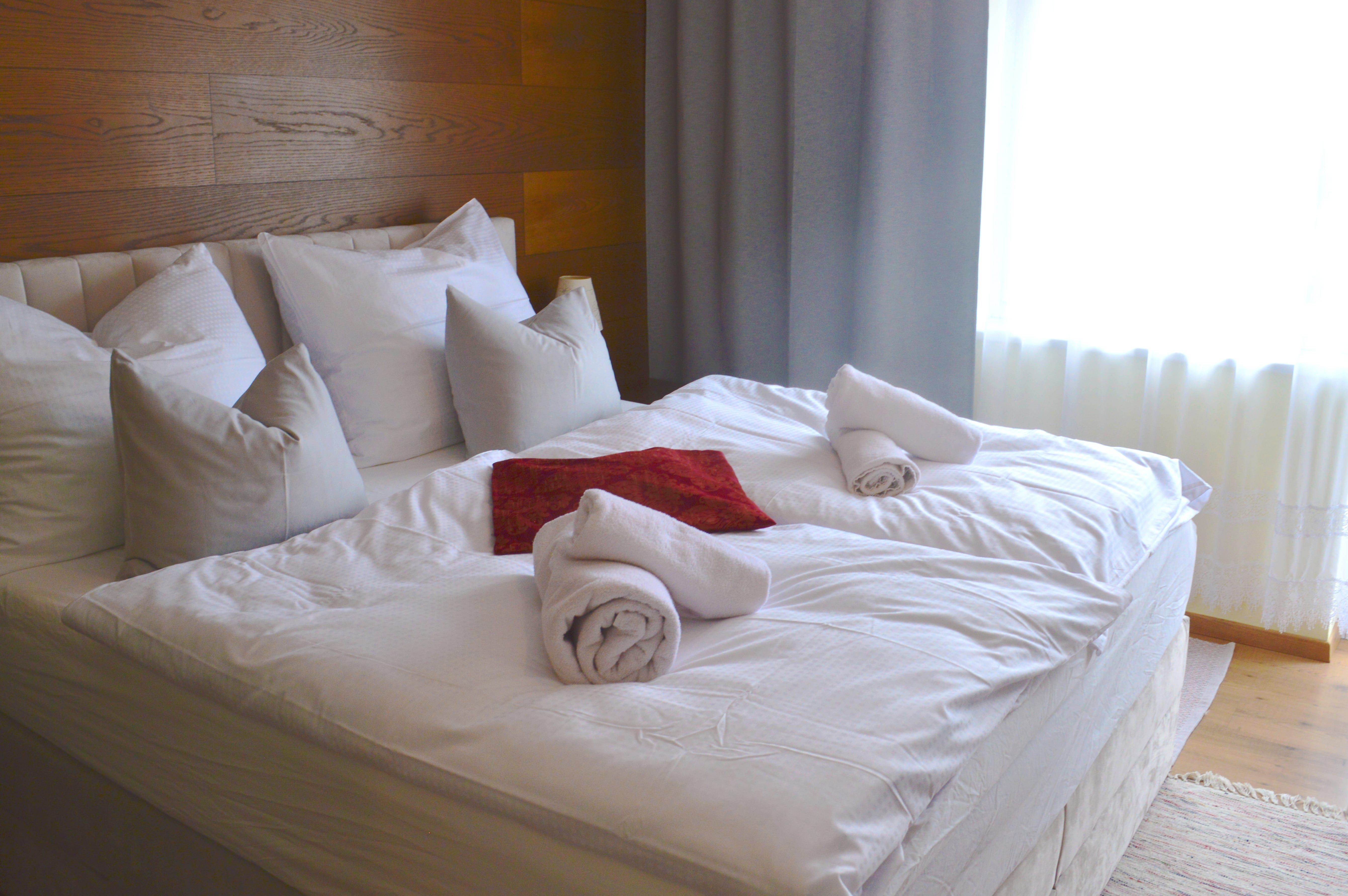 Ein gemütliches Doppelbett mit weißen Kissen und Handtüchern, dekoriert mit einem roten Tuch, in einem hellen Zimmer mit Holzwand und Vorhängen.