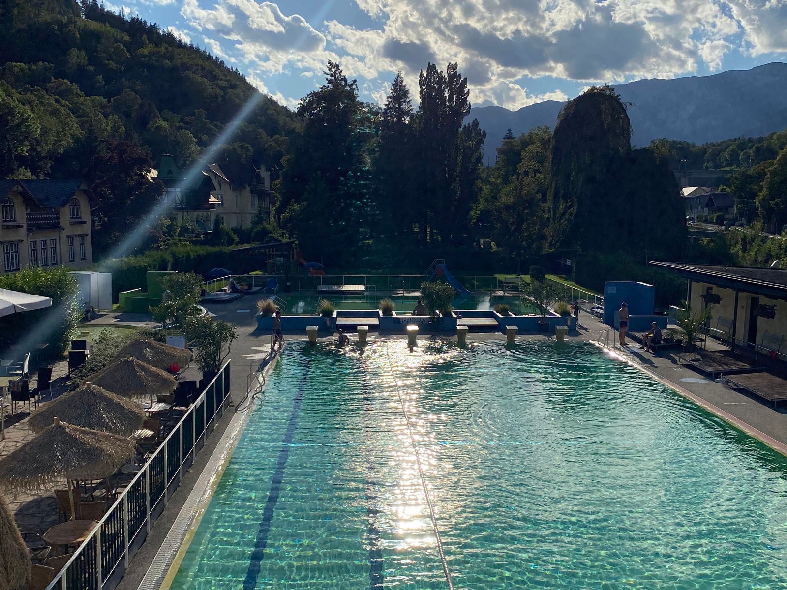 Ein Freibad mit klarem Wasser, umgeben von Bäumen und Bergen im Hintergrund.