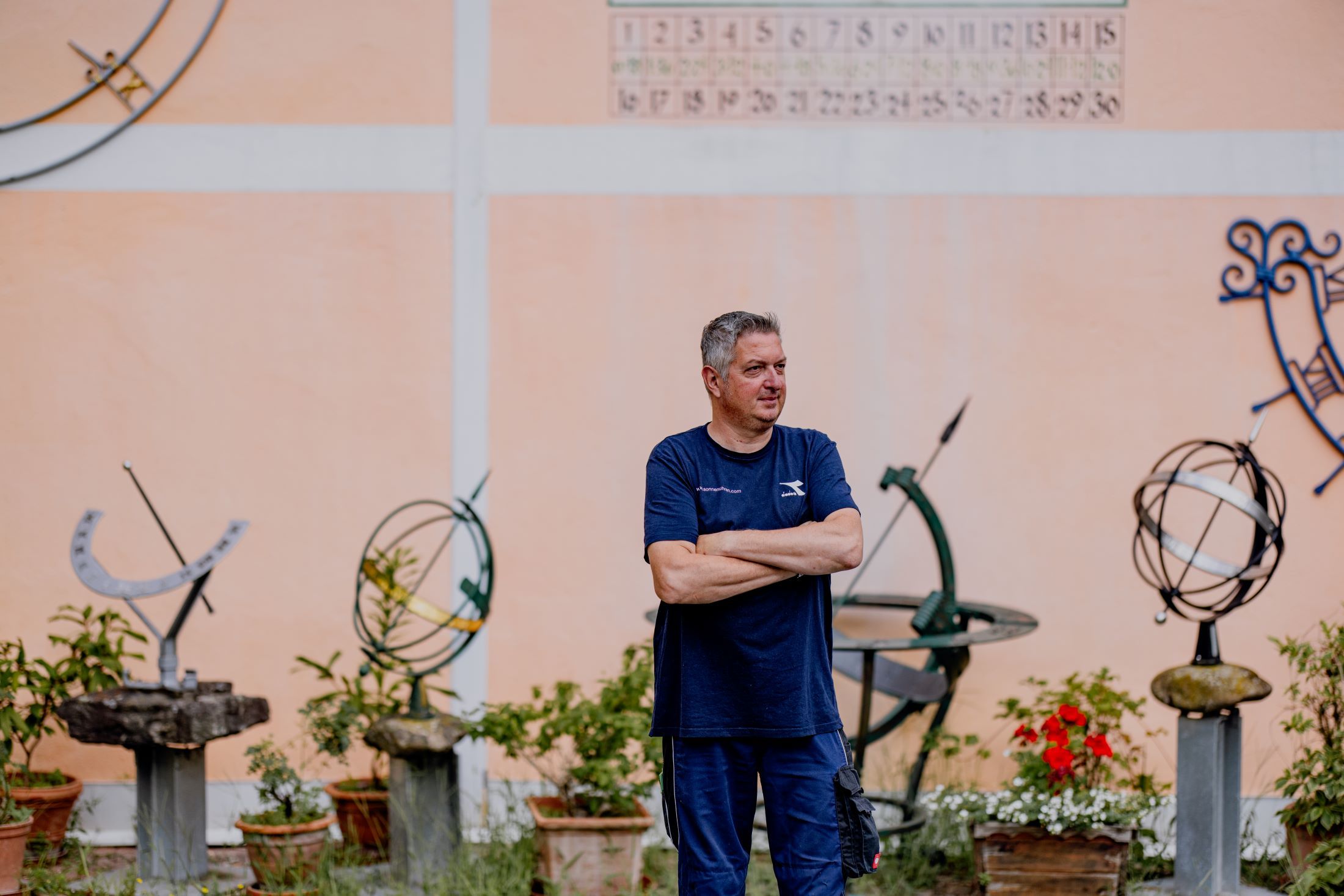 Ein Mann steht mit verschränkten Armen vor einer Wand mit Sonnenuhren und Pflanzen.