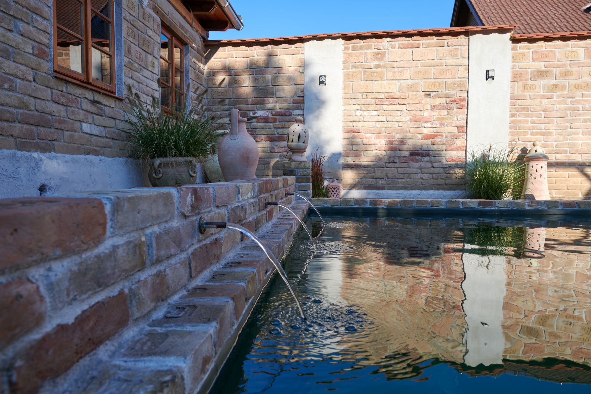Backsteinmauer mit Wasserspeiern und Pflanzen am Rand eines Pools.