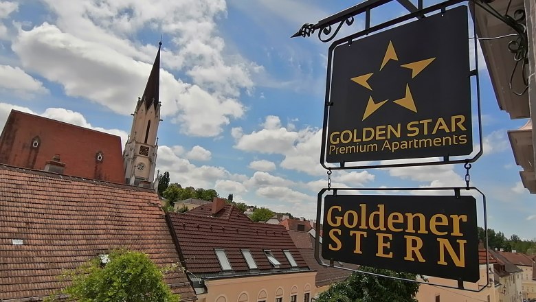 Blick auf ein Schild mit der Aufschrift 'Golden Star Premium Apartments' und 'Goldener Stern' vor einem Kirchturm und Dächern.