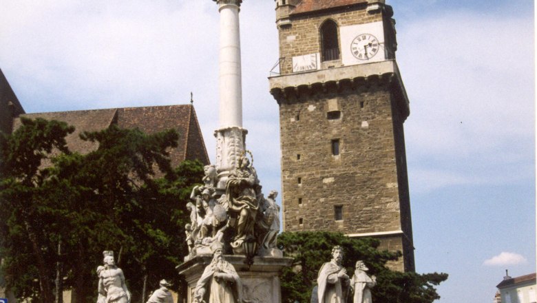 Historischer Wehrturm mit Uhr und barocker S&auml;ule im Vordergrund.