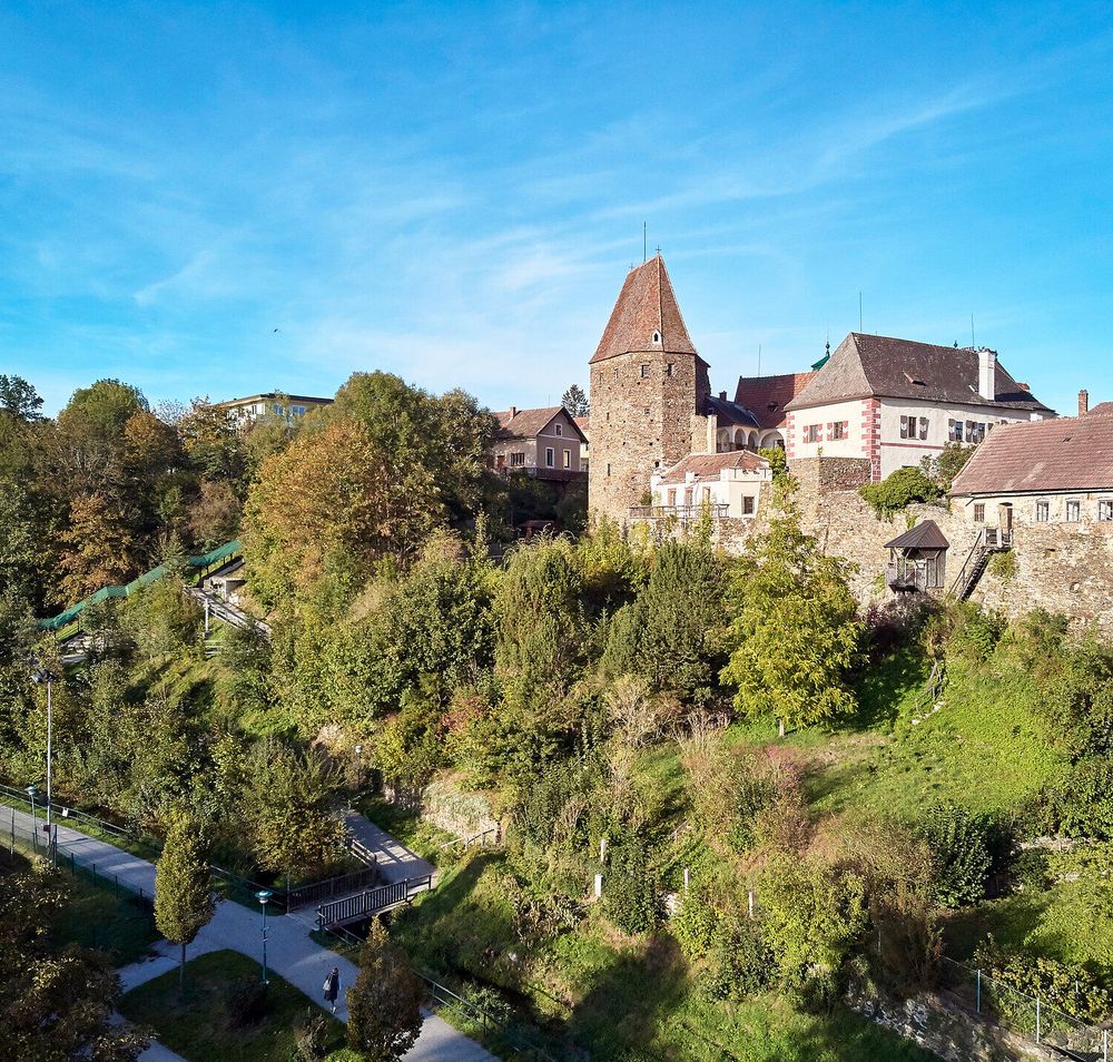 Die sanften Hügel umgeben die charmante Stadt, während die historische Stadtmauer majestätisch in den Himmel ragt. Bunte Bäume und üppiges Grün verleihen der Umgebung eine einladende Atmosphäre, die zum Verweilen und Entdecken einlädt.