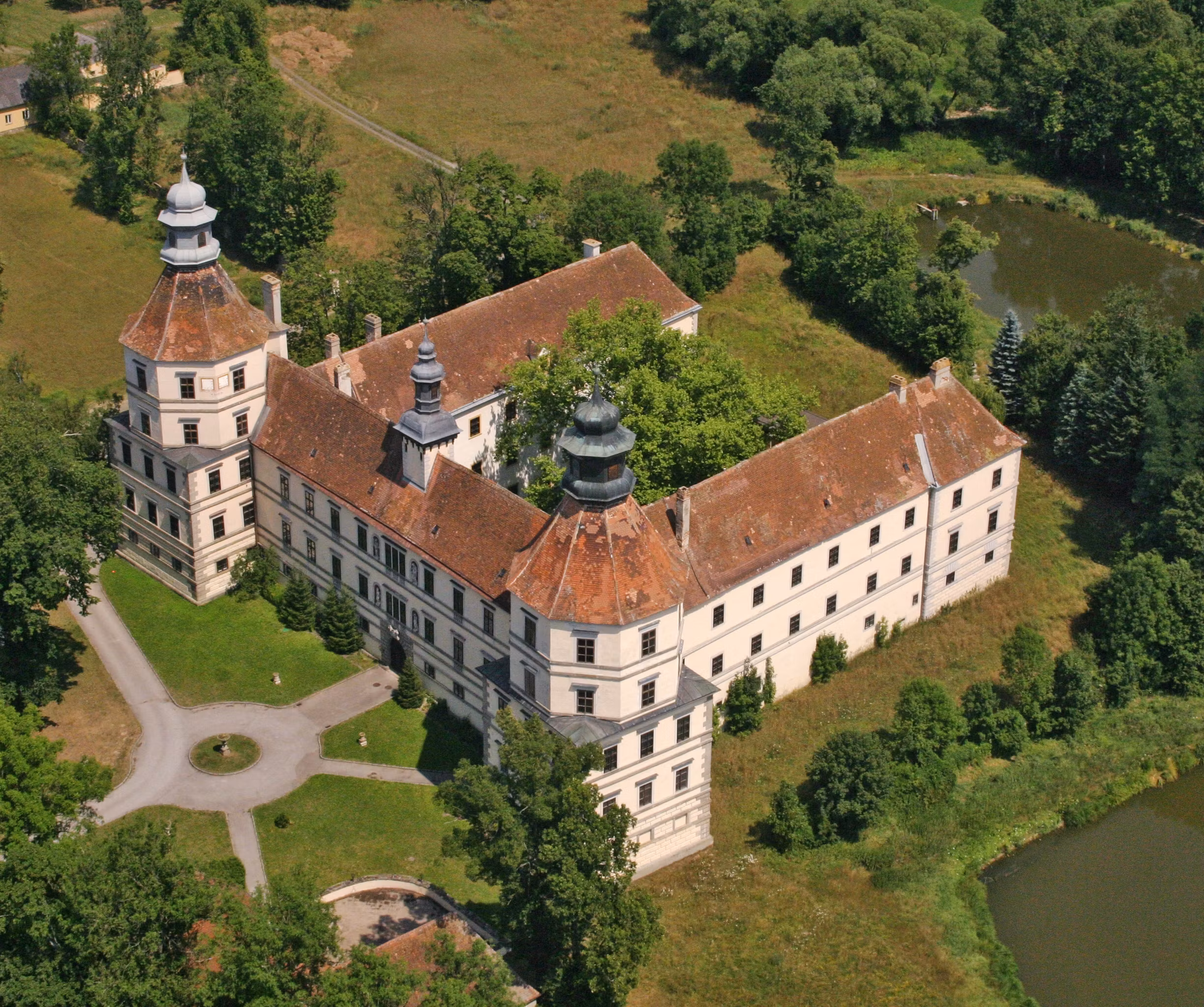 Luftaufnahme eines historischen Schlosses mit zwei Türmen und einem Innenhof, umgeben von Bäumen und einem Teich.