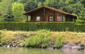 Ein Holzferienhaus mit rotem Dach, umgeben von B&auml;umen und Str&auml;uchern, steht am Ufer eines Gew&auml;ssers.