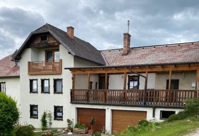 Ein zweistöckiges Haus mit Balkon und Garage, umgeben von grüner Landschaft und einem bewölkten Himmel.