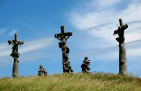 Drei Kreuze mit Figuren auf einem H&uuml;gel unter blauem Himmel.