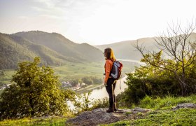 Eine Frau mit Rucksack steht auf einem Hügel und blickt auf eine Flusslandschaft mit Bergen im Hintergrund.