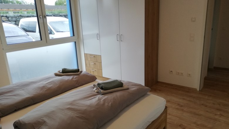 Schlafzimmer im Erdgeschoss mit Holzboden, -bett, wei&szlig;em Schrank und gro&szlig;em Fenster.