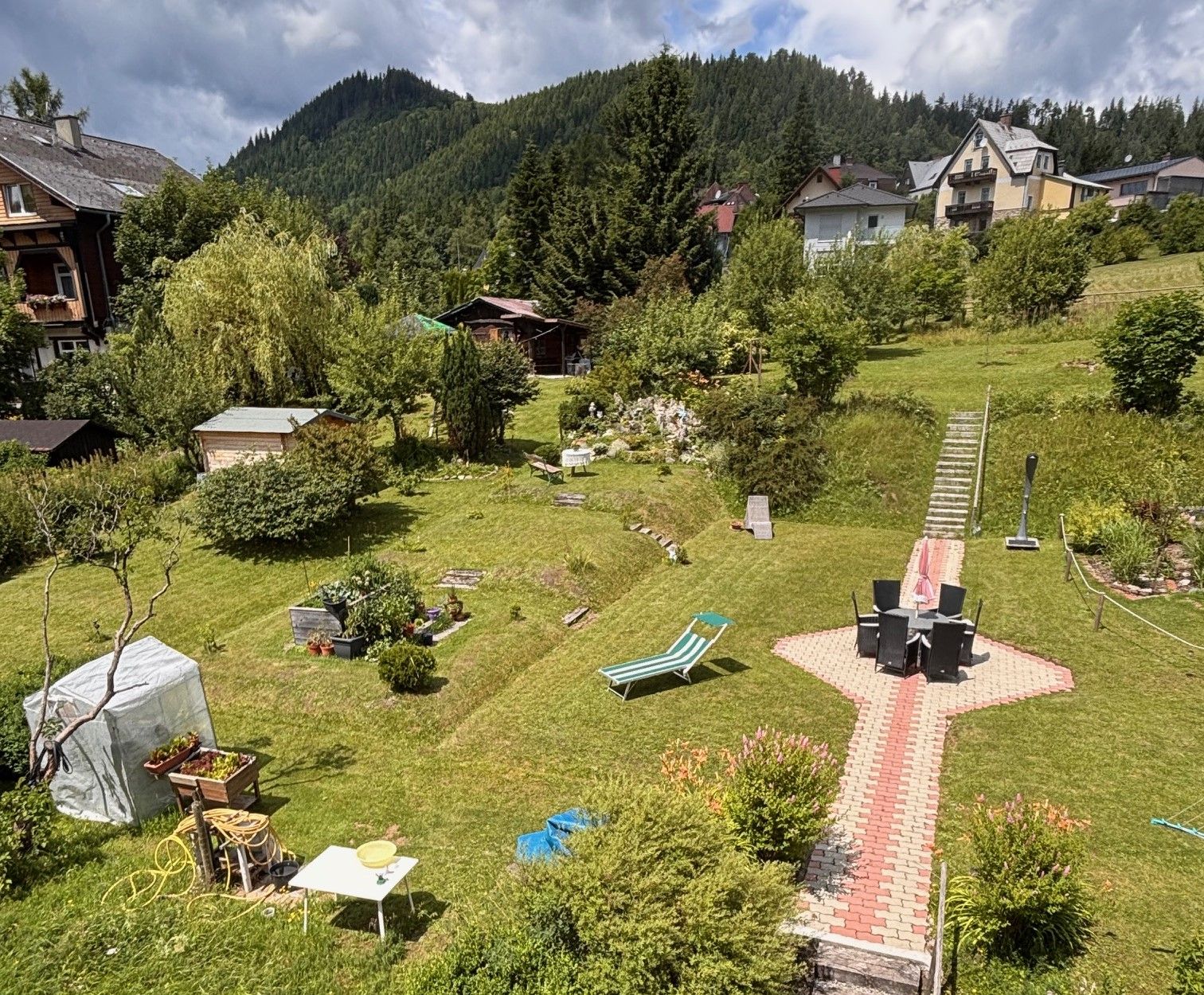 Ein gepflegter Garten mit Rasen, Gartenmöbeln und einem Weg aus Pflastersteinen. Im Hintergrund sind Häuser und bewaldete Hügel zu sehen.