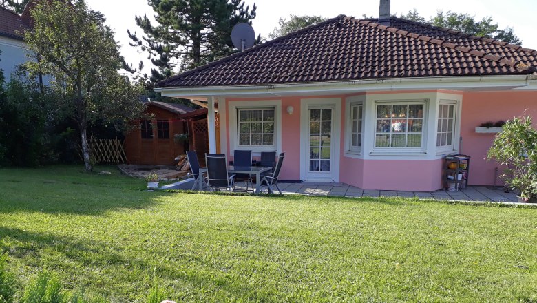 Ein rosa Haus mit Garten, Terrasse und Gartenm&ouml;beln, umgeben von B&auml;umen und Blumen.