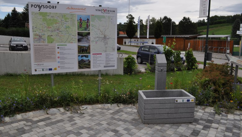 Ein Trinkbrunnen neben einer Informationstafel in Poysdorf, umgeben von Blumen und parkenden Autos.