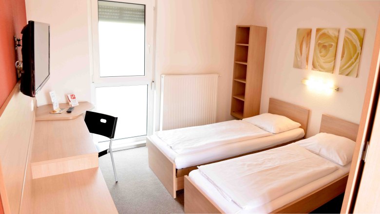 Ein einfaches Motelzimmer mit zwei Einzelbetten, einem Schreibtisch, einem Stuhl und einem Fernseher an der Wand.