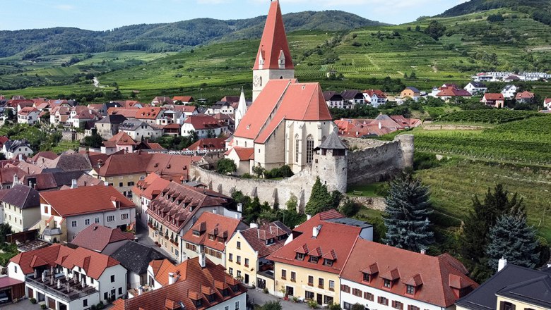 Luftaufnahme eines Dorfes mit Kirche und Weinbergen im Hintergrund.