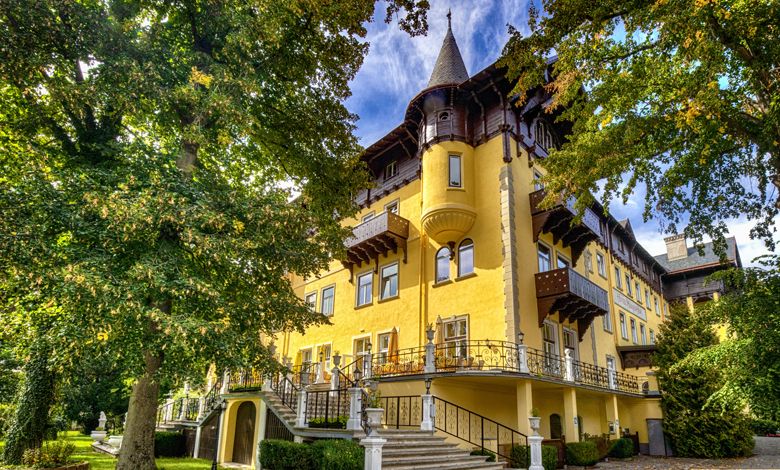 Historisches gelbes Gebäude mit Türmen und Verzierungen, umgeben von Bäumen.