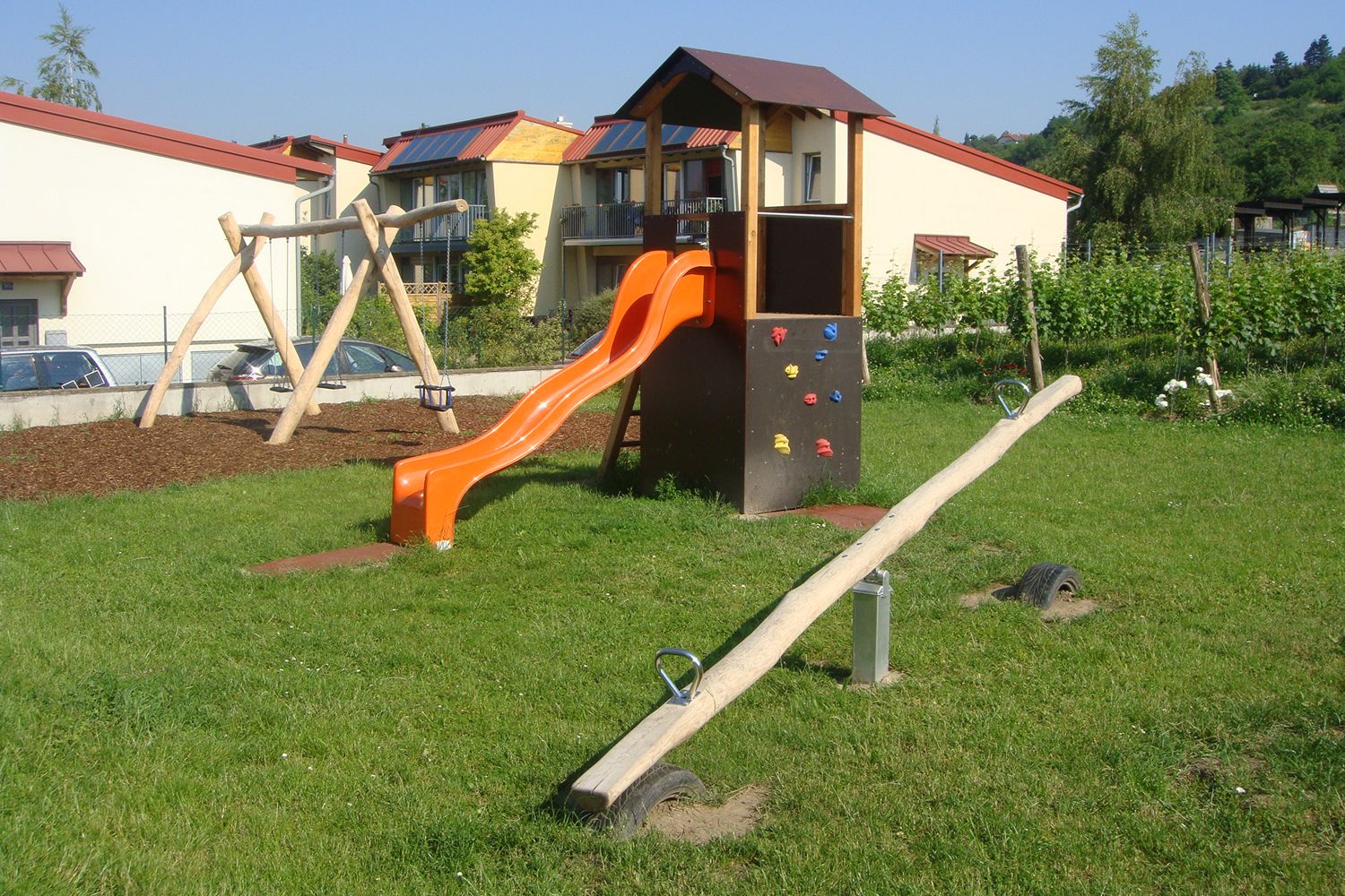 Ein Kinderspielplatz mit Rutsche, Schaukel und Wippe auf einer Wiese vor Wohnhäusern.