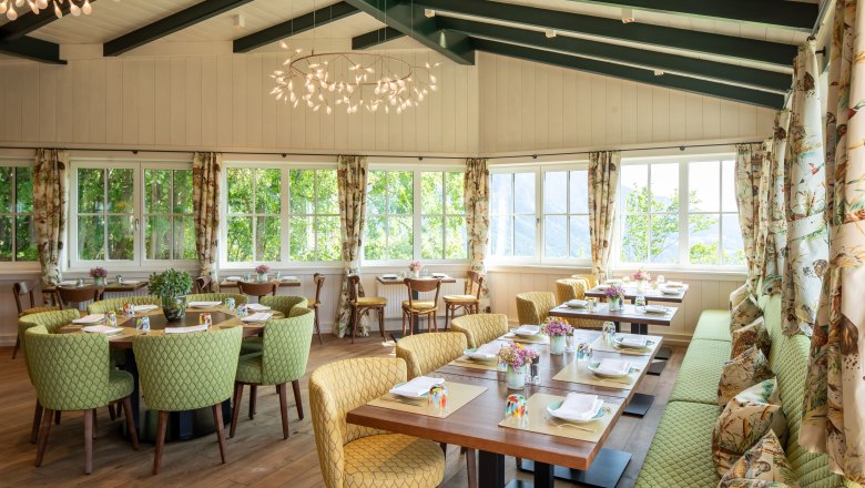 Ein heller Wintergarten mit gedeckten Tischen, gr&uuml;nen St&uuml;hlen und gro&szlig;en Fenstern.