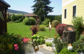 Ein gepflegter Garten mit Blumen, Sträuchern und einem gelben Haus im Hintergrund.