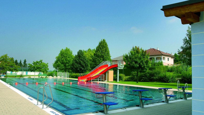 Ein Freibad mit einem Schwimmbecken, einer roten Rutsche und Startbl&ouml;cken. Im Hintergrund sind B&auml;ume und ein Wohnhaus zu sehen.